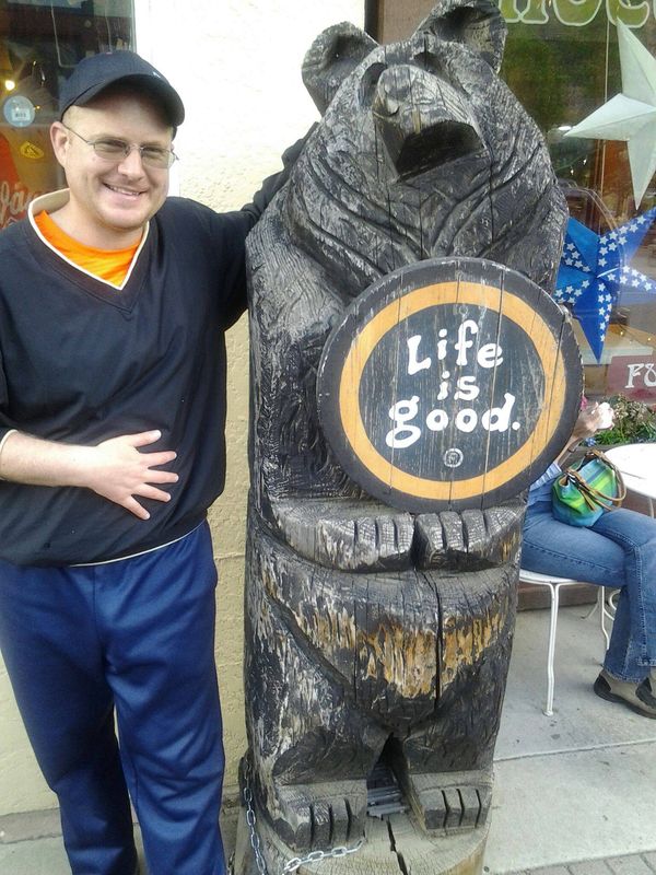 Robert Jenkins standing next to a carved wooden bear holding a Life is good sign in downtown Idaho Springs, Colorado