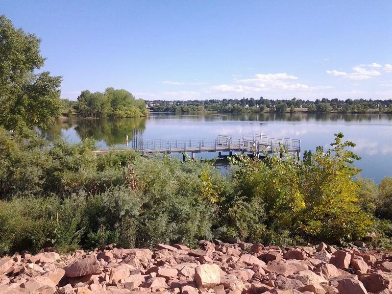 Quincy Reservoir in Aurora, Colorado, with a fishing pier, trees along the shore and red rocks under a blue sky